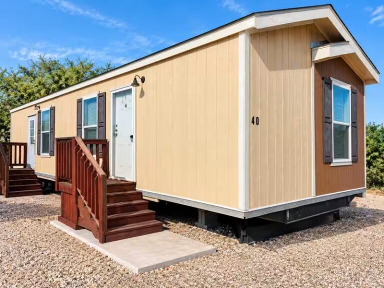 Mobile home with tan siding and wooden stairs, marked "40," on gravel lot under blue sky.