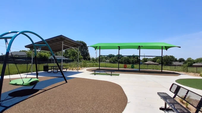 Children's playground with green canopy shades, swing sets, benches, and picnic pavilion in grassy park.