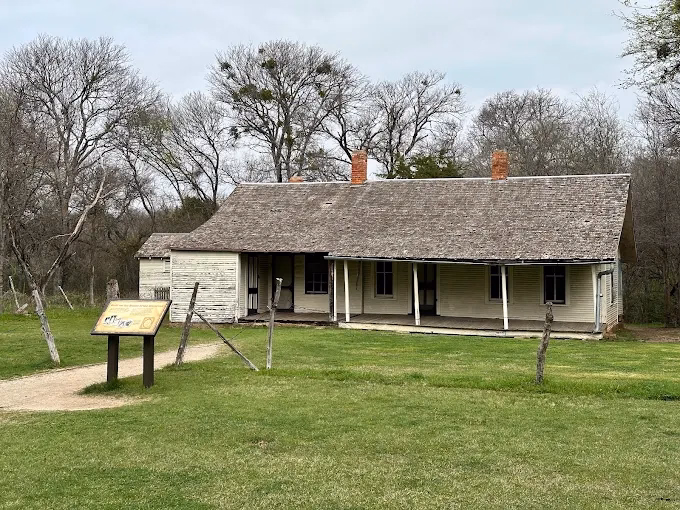 Historic white wooden cottage with front porch surrounded by bare winter trees and grassy lawn.