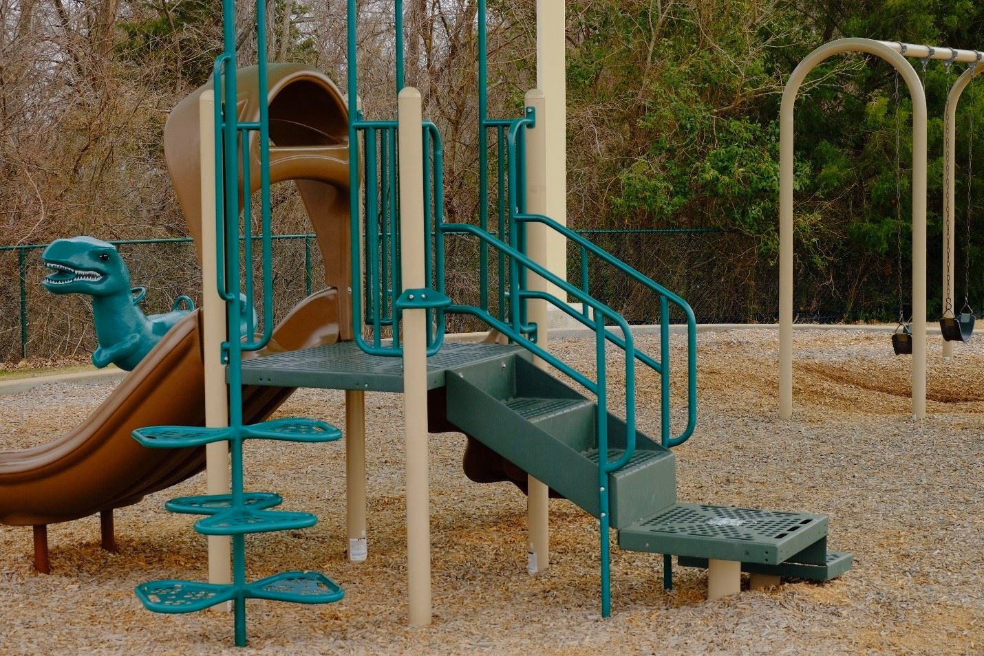 Green playground structure with blue tube slide at a park surrounded by trees.