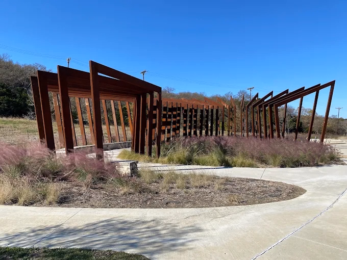 A rustic wooden pergola and slat fencing line a curved concrete walkway bordered by tall ornamental grasses.