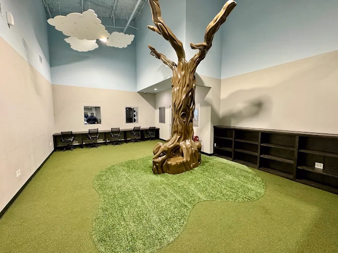 Artificial tree standing indoors in a gallery-like room with light walls, dark shelving, and green flooring.