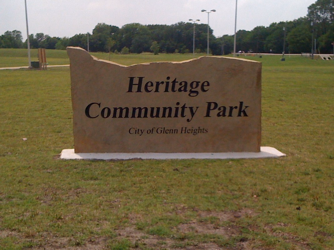 Stone welcome sign in a park field that reads Heritage Community Park, with green trees and lawn behind it.