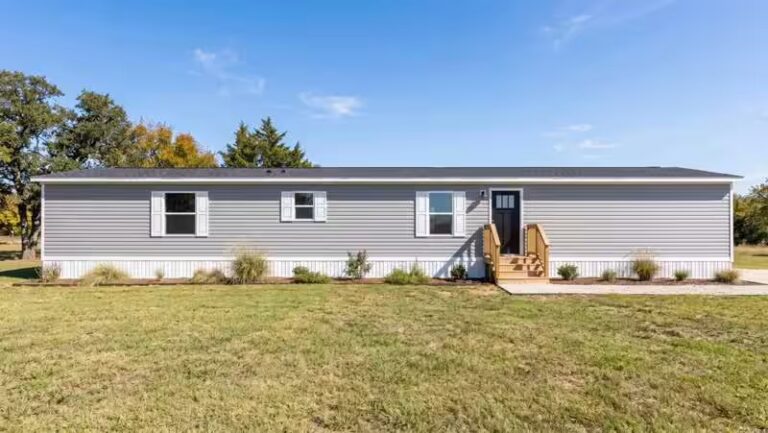 Gray mobile home with white shutters, wooden front steps, and lawn under blue sky.