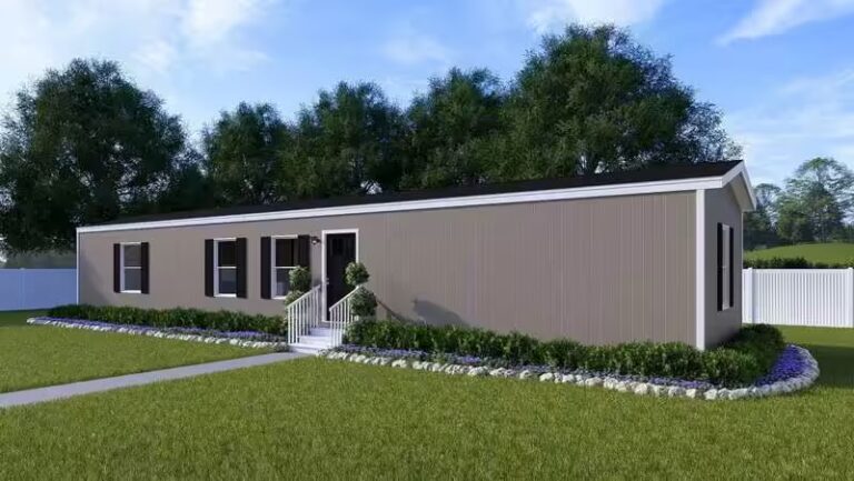 Single-story tan manufactured home with black shutters, porch, and landscaped yard before trees.