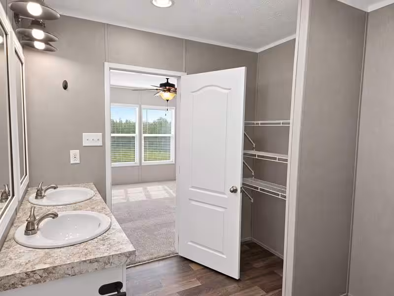 Double-sink vanity with granite counter, open white door to bedroom with ceiling fan and windows.