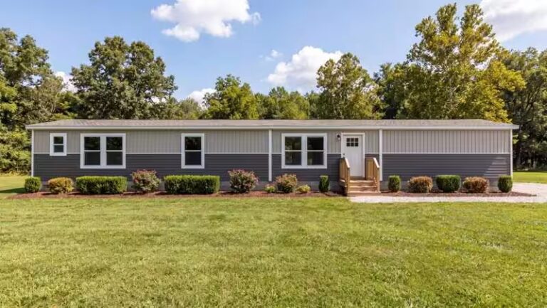 Gray single-wide mobile home with white trim, small porch, and green lawn surrounded by trees.