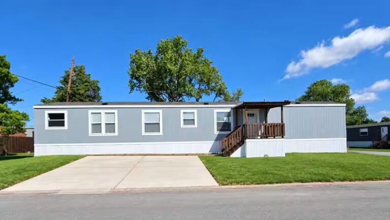 Gray mobile home with wooden porch and concrete driveway under a blue sky with trees behind.