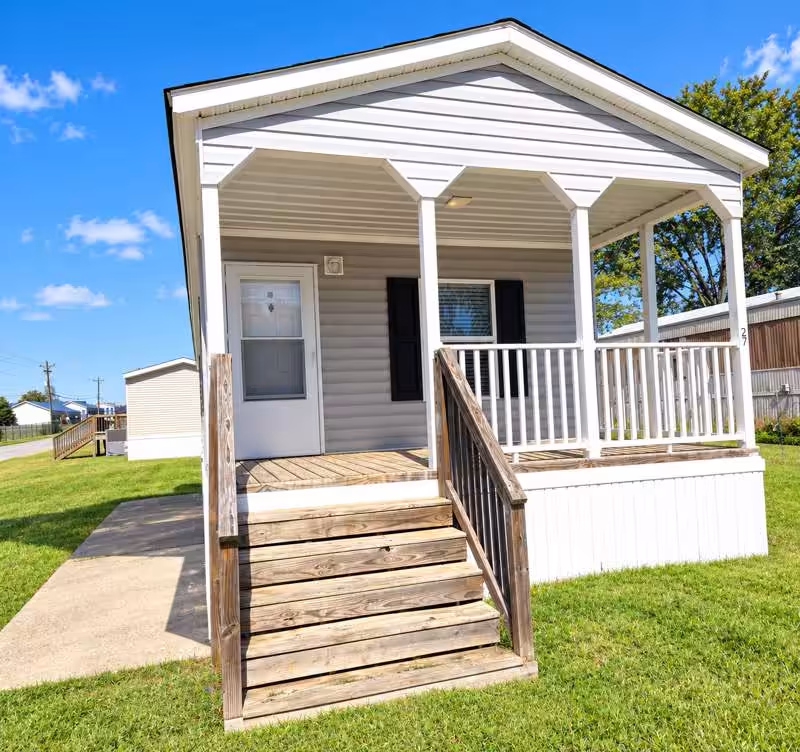 White mobile home with covered porch, white railings, and wooden steps on a grassy lawn under blue sky.