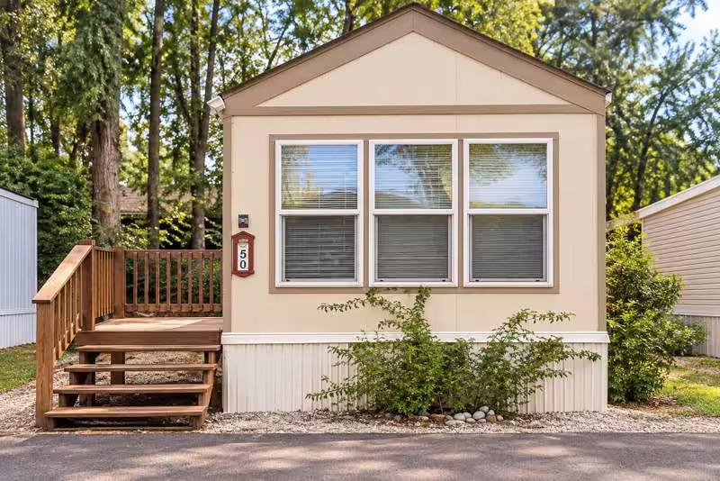 Small beige mobile home with wooden porch stairs, number 50 sign, surrounded by trees.