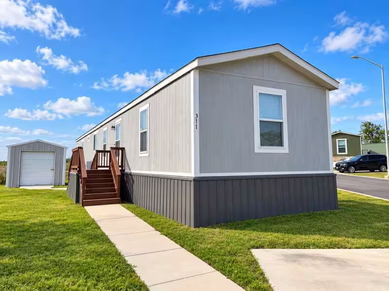 Gray mobile home with wooden stairs on green lawn under blue sky, number 311 on the wall.